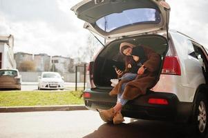 Young mother and child sitting in the trunk of a car and looking at mobile phone. Safety driving concept. photo