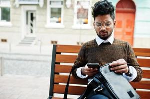 Portrait of young stylish indian man model pose in street, sitting on bench with handbag and smartphone. photo