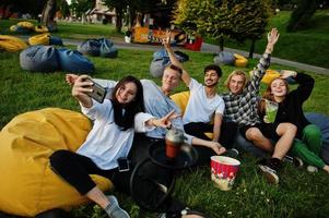 Young multi ethnic group of people watching movie at poof in open air cinema and making selfie on phone. photo