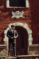 Portrait of retro 1920s english arabian business man wearing dark suit, tie and flat cap standing against old brick house 1898 year. photo