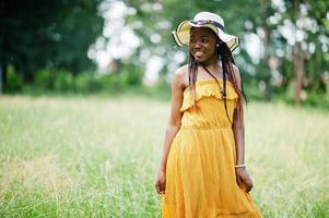 retrato de una hermosa mujer afroamericana de 20 años vestida con vestido amarillo y sombrero de verano posando en la hierba verde en el parque. foto