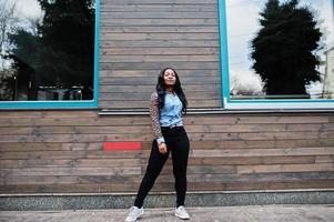 Hipster african american girl wearing jeans shirt with leopard sleeves posing at street against wooden house with windows. photo