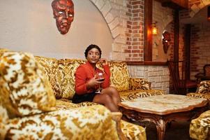 African american woman,sitting on the couch,  retro hairstyle, wear orange jacket and leather skirt posing at restaurant with glass of wine. photo