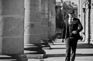 Portrait of retro 1920s english arabian business man wearing dark suit, tie and flat cap near old columns. photo