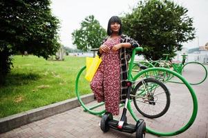 hermosa mujer afroamericana de pie cerca de segway o hoverboard y bicicleta. chica negra con símbolo de reciclaje de bolsas ecológicas de tela amarilla. foto