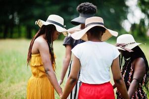 Group of four gorgeous african american womans wear summer hat holding hands and praying at green grass in park. photo