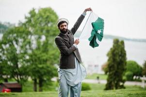 El hombre pakistaní de barba usa sombrero pakol y chaqueta sostiene la bandera de pakistán. foto