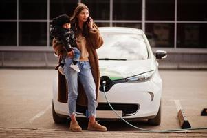 madre joven con un niño cargando un auto eléctrico en la gasolinera eléctrica y hablando por teléfono móvil. foto