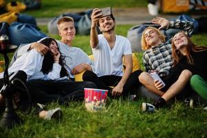 Young multi ethnic group of people watching movie at poof in open air cinema and making selfie on phone. photo