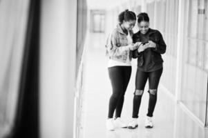 dos amigas africanas con chaqueta de jeans usando teléfonos móviles juntos en el interior. foto