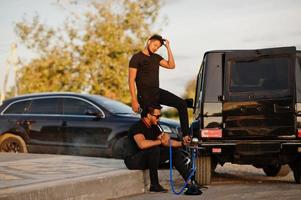 Two asian brothers man wear on all black posed near suv cars and smoke hookah. photo