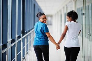 Back of two african woman friends in t-shirts walking halding hands indoor together. photo