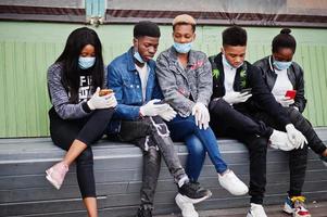 Group of african teenagers friends sitting with phones, wearing medical masks protect from infections and diseases coronavirus virus quarantine. photo