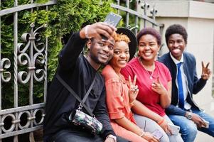 cuatro amigos africanos divirtiéndose al aire libre. dos chicas negras con chicos con teléfonos móviles hacen selfie. foto