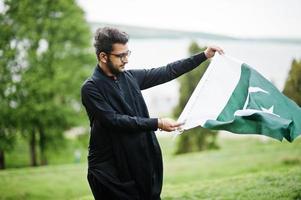 pakistano bangladesh el hombre usa ropa tradicional negra y anteojos posan al aire libre, sosteniendo la bandera de pakistán. foto