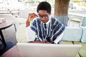 African man in traditional clothes and glasses sitting behind laptop at outdoor caffe. photo