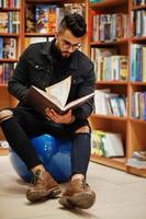 estudiante árabe alto e inteligente, vestido con chaqueta de jeans negros y anteojos, sentado en la biblioteca y leyendo un libro en las manos. foto