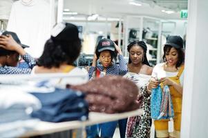 Three african woman choosing clothes at store. Shopping day. They buying caps and scarfs and looking at mirror. photo