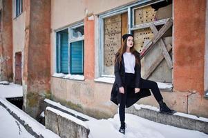Fashionable long legs brunette model in long black cloak posed outdoor at winter day against old grunge wall with broken windows. photo