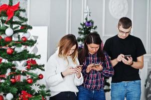 grupo de amigos como dos chicas y un hombre mirando teléfonos móviles contra el árbol de navidad en el estudio. foto