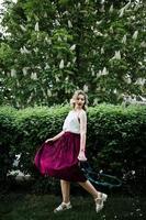 Fashionable and beautiful blonde model girl in stylish red velvet velour skirt, white blouse, posed at park with phone and earphones. photo