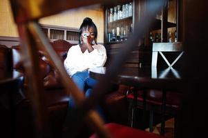 Stylish african american women in white blouse and blue jeans posed at cafe with newspaper. photo