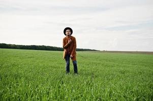 hombre elegante con gafas, chaqueta marrón y sombrero posado en campo verde. foto