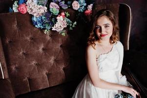 Pretty young gilrl in white dress sitting on brown vintage sofa with flowers. photo