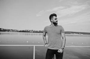 apuesto modelo de hombre de barba árabe alto en camisa desnuda posada al aire libre en el muelle del lago. chico árabe de moda. foto