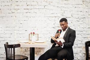 Strong powerful african american man in black suit sitting at cafe, read book and sniff flower in test tube. photo