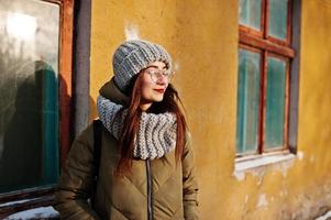 Portrait of brunette girl in gray scarf and hat, glasses at cold weather with sunshine against orange wall of old house. photo