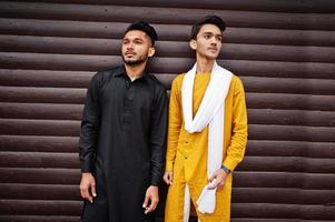 Two indian stylish mans friends in traditional clothes posed outdoor against wooden background. photo