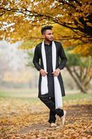 Indian stylish man in black traditional clothes with white scarf posed outdoor against yellow autumn leaves tree. photo