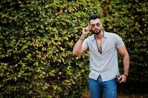 Fashionable tall arab beard man wear on shirt, jeans and sunglasses posed on park against greenery. photo