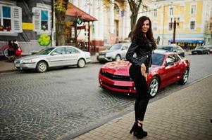 joven mujer rizada y sexy en chaqueta de cuero contra muscle car rojo en la calle. foto