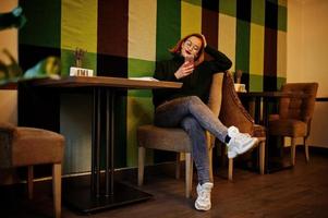 joven alegre y hermosa mujer pelirroja con gafas, suéter verde de lana caliente, usando su teléfono, mientras se sienta en su lugar de trabajo en el café. foto