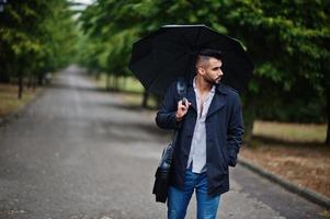 hombre de barba árabe alto de moda vestido con abrigo negro con paraguas y estuche posado en el día de la lluvia. foto