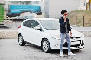 South asian man or indian male wear red eyeglasses stand near his white transportation with mobile phone. photo