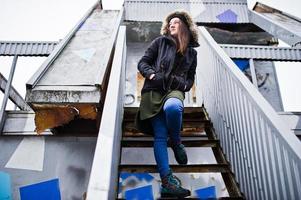 ropa de niña con sudadera verde larga, jeans y sombreros negros en un lugar industrial. foto