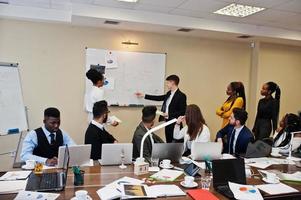 Mixed race business coach presenting report standing near whiteboard pointing on sales statistic shown on diagram and chart teach diverse company members gathered together in conference room. photo