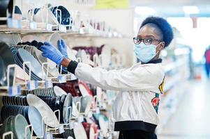 African woman wearing disposable medical mask and gloves shopping in supermarket during coronavirus pandemia outbreak. Black female choose bowl at epidemic time. photo