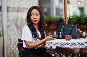 elegante mujer de negocios afroamericana sentada en un café al aire libre con teléfono móvil. foto