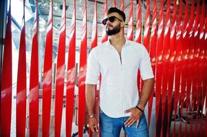 Stylish tall arabian man model in white shirt, jeans and sunglasses posed at street of city. Beard attractive arab guy against red wall. photo