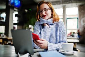Cheerful young beautiful redhaired woman in glasses using her phone, touchpad and notebook while sitting at her working place on cafe with cup of coffee. photo