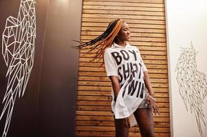 hermosa mujer africana con ropa informal elegante posando contra la pared de alas de ángel. ella moviendo sus rastas de pelo. foto