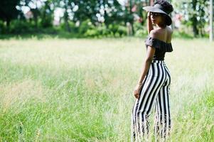 Portrait of gorgeous african american woman 20s in wear in black and white stripe pants and summer hat posing at green grass in park. photo