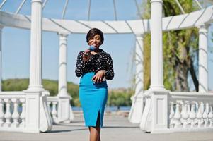 Stylish african american model girl TV presenter with microphone in glasses, blue skirt and black blouse posed outdoor against white stone arch with mobile phone. photo