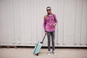 African american man in checkered shirt, sunglasses and earphones with suitcase and backpack. Black man traveler against wall. photo