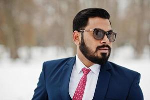 primer plano retrato de elegante hombre de negocios de barba india en traje y gafas de sol posado en el día de invierno al aire libre. foto