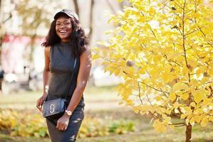 Stylish african american girl in gray tunic, crossbody bag and cap posed at sunny autumn day against yellow leaves. Africa model woman. photo